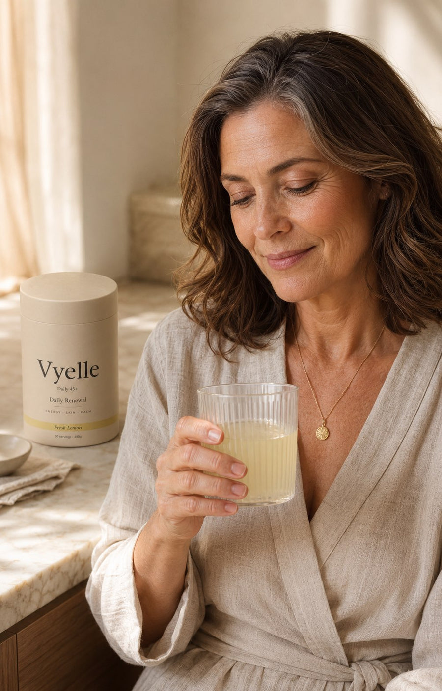 Woman in soft morning light holding a glass of Vyelle Daily Renewal.